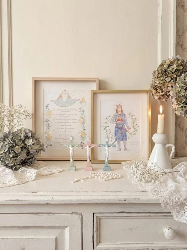 Composition décorative de style shabby chic sur une commode en bois blanc vieilli, présentant un cadre doré avec l'illustration de Saint Louis, un cadre en bois clair avec les Dix Commandements, trois crucifix pastels (vert menthe, rose, bleu clair) avec un corpus blanc, un chapelet blanc, une bougie allumée dans un bougeoir en céramique, des bouquets d'hortensias séchés et de gypsophile, et de la dentelle blanche.