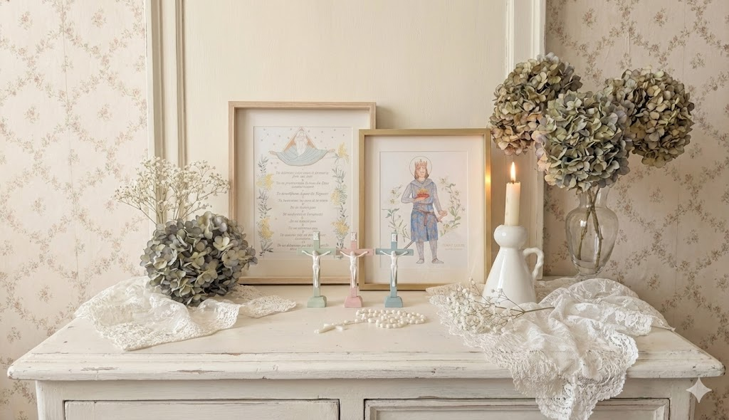 Bannière _Composition décorative de style shabby chic sur une commode en bois blanc vieilli, présentant un cadre doré avec l'illustration de Saint Louis, un cadre en bois clair avec les Dix Commandements, trois crucifix pastels (vert menthe, rose, bleu clair) avec un corpus blanc, un chapelet blanc, une bougie allumée dans un bougeoir en céramique, des bouquets d'hortensias séchés et de gypsophile, et de la dentelle blanche.