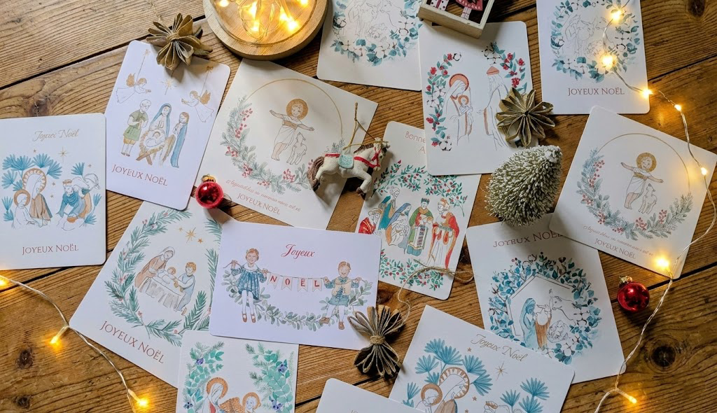 Composition à plat de cartes de Noël françaises traditionnelles et illustrées, étalées sur une table en bois. On y voit des scènes de la Nativité, des enfants tenant une bannière 'Joyeux Noël', une guirlande lumineuse allumée, un petit cheval de bois et des étoiles d'anis.
